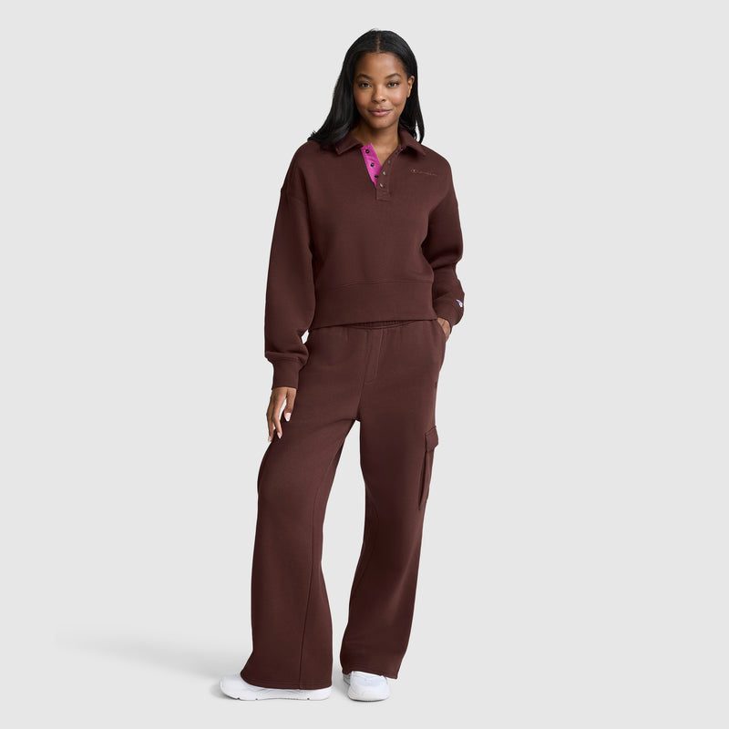 A woman stands smiling, wearing a matching dark brown pullover and Champion Fleece Cargo Training Pant, paired with white sneakers against a plain light gray background.