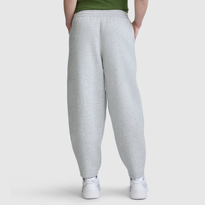 A person is wearing the Champion Barrel Leg Fleece Pant in light gray, paired with white sneakers and a green top. They are standing with hands behind their back, facing away from the camera against a plain background.