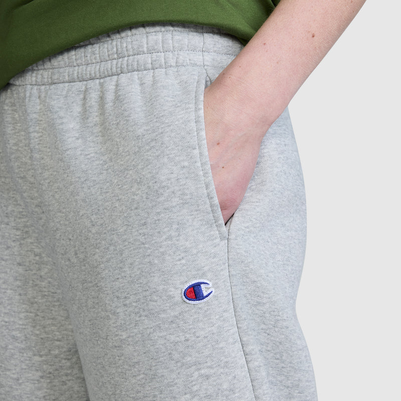 A person wearing Champion Barrel Leg Fleece Pant in light gray with the Champion logo visible on the pants and one hand in the pocket. They are also wearing a green shirt.