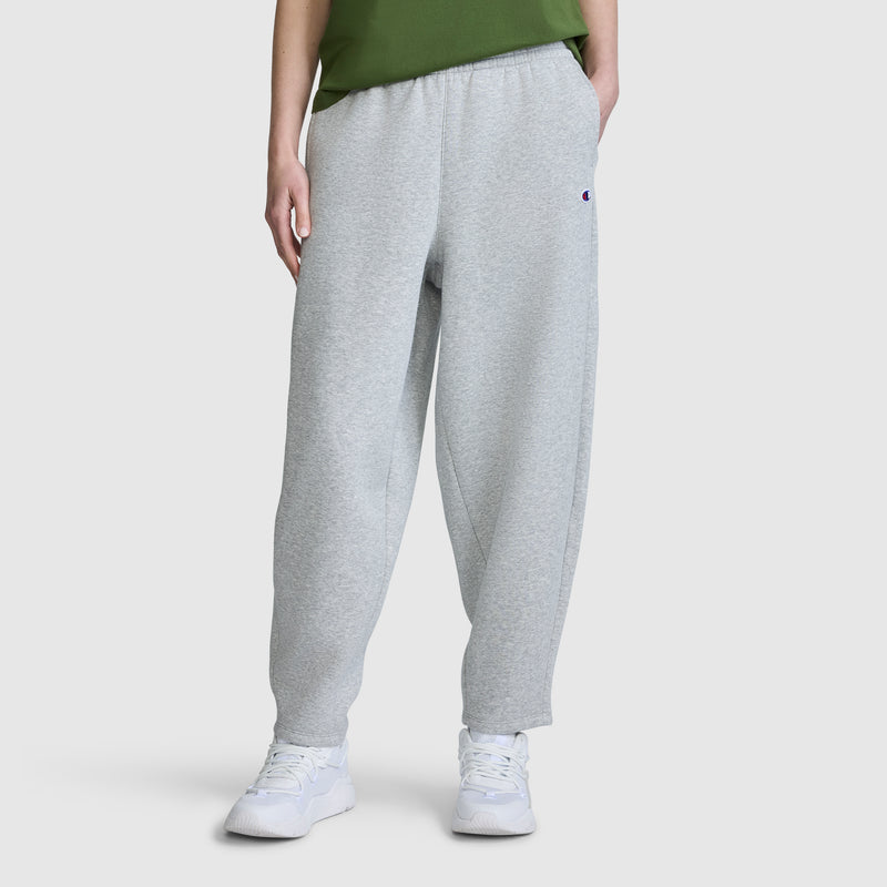 A person is shown from the waist down, wearing the Champion Barrel Leg Fleece Pant in light gray, paired with a green shirt and white sneakers, against a plain white background. The relaxed fit of these Champion women’s pants is prominently displayed.