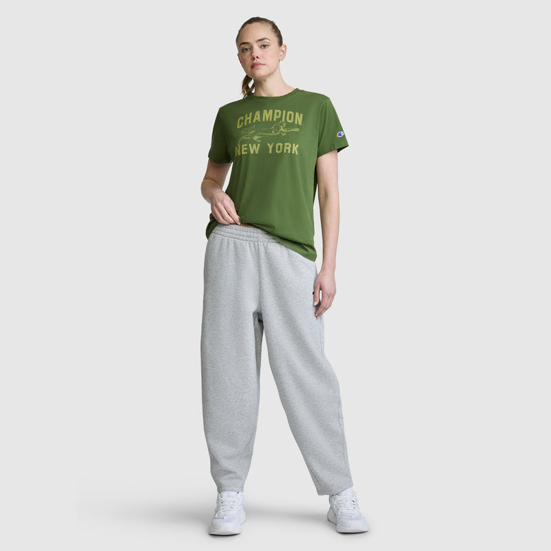 A woman stands against a plain white background wearing a green Champion New York t-shirt, Champion Barrel Leg Fleece Pant in light gray, and white sneakers.