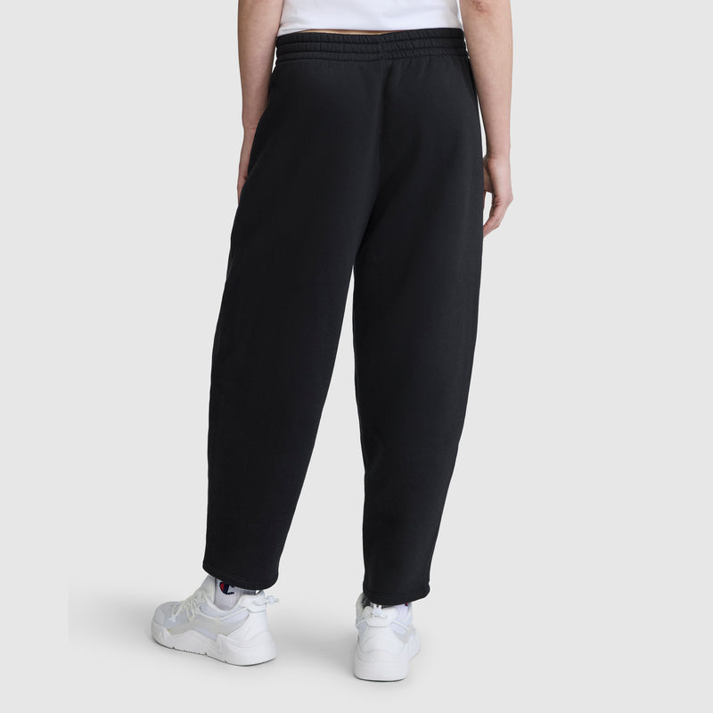 A person stands with their back to the camera against a plain light background, wearing Champion Barrel Leg Fleece Pants made from midweight cotton-poly fleece, paired with a white T-shirt and white sneakers.