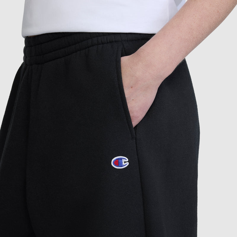 A person is seen wearing Champion's Barrel Leg Fleece Pant in black, with one hand tucked into the pocket. Only the lower torso and part of an arm are visible, showcasing the embroidered Champion logo on these midweight cotton-poly sweatpants.