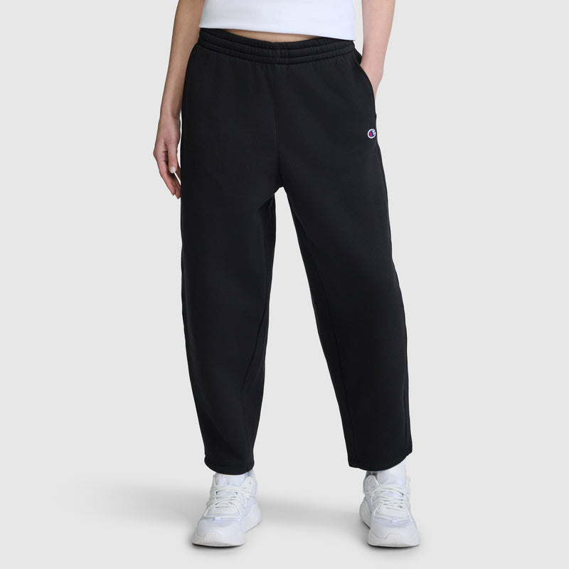 A person is wearing the Champion Barrel Leg Fleece Pant, crafted from midweight cotton-poly fleece in black, paired with a white cropped top and white sneakers, standing against a plain light gray background.