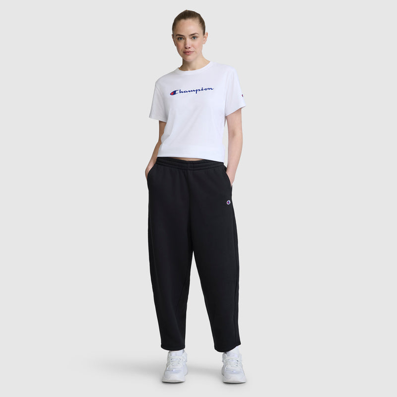 A woman is standing with her hands in the pockets of loose black Barrel Leg Fleece Pant by Champion, paired with a white Champion T-shirt and white sneakers, set against a plain light gray background.