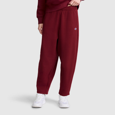 A person is shown from the waist down wearing Champion Barrel Leg Fleece Pants in maroon, paired with a matching sweatshirt and white sneakers, standing against a white background.