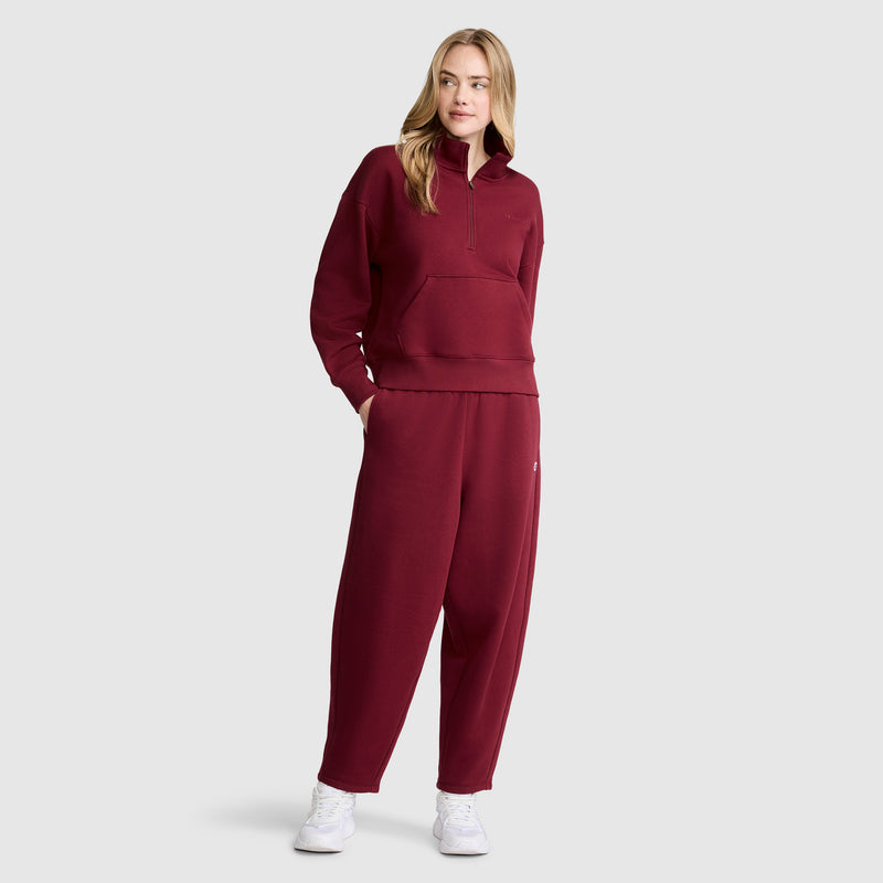A woman stands against a gray background, wearing the Champion Barrel Leg Fleece Pant with a matching zip-up top and loose, comfortable pants in burgundy, paired with white sneakers. She has long blonde hair and a relaxed expression.