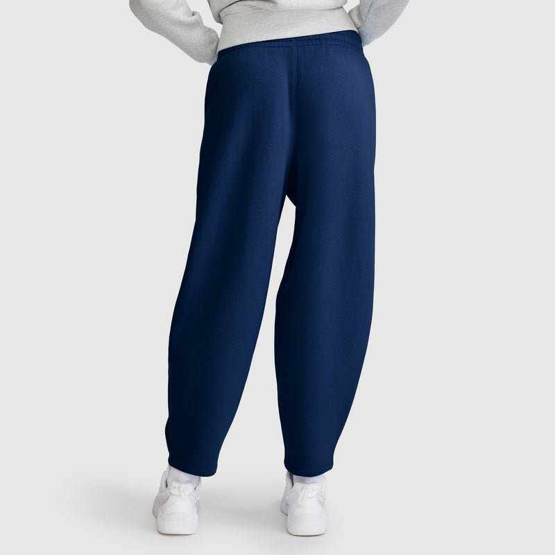 A person is shown from the back against a plain light background wearing the Champion Barrel Leg Fleece Pant in navy blue, made from soft midweight fleece, paired with white sneakers and a light gray sweatshirt.