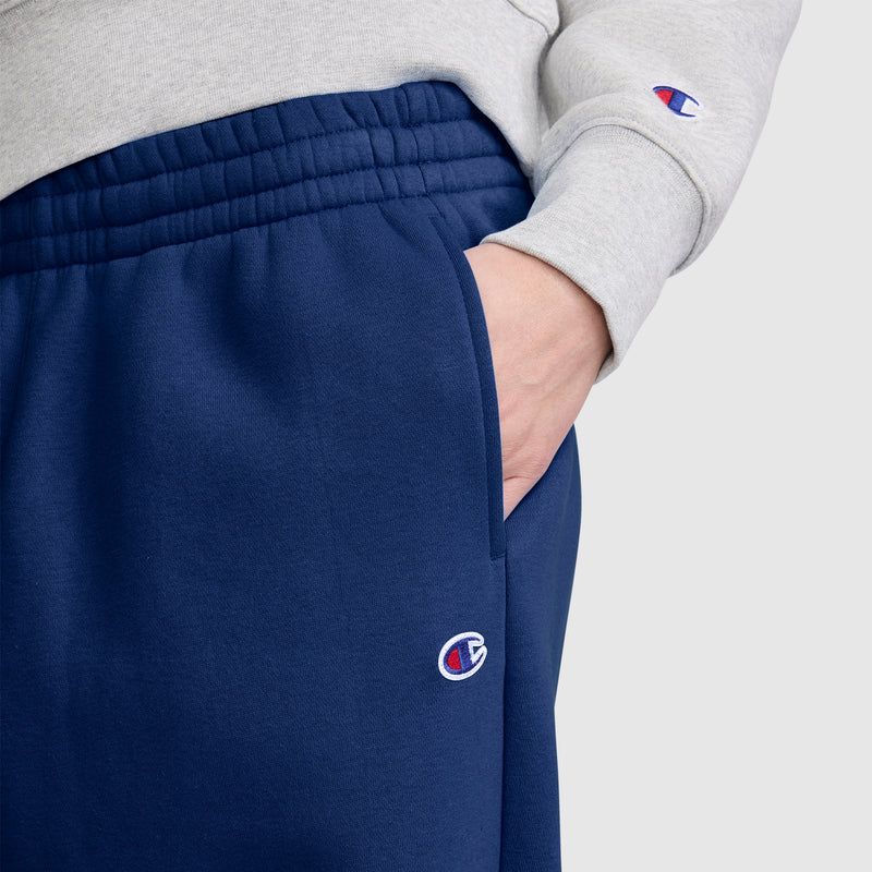 A person wearing a gray sweatshirt and blue Champion Barrel Leg Fleece Pants has their right hand in their pocket. The image highlights the waist and hip area, showcasing the midweight fleece fabric and the relaxed barrel leg style from Champion.