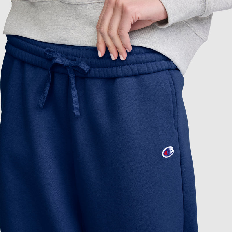 A person wearing a light gray sweatshirt and navy blue Champion Barrel Leg Fleece Pants adjusts the waistband with one hand. The midweight fleece offers cozy warmth, blending comfort and style in this casual Champion look.