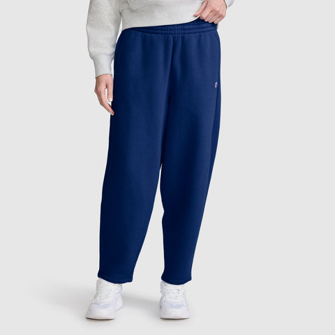 Dressed in Champion Barrel Leg Fleece Pant in navy blue, paired with a light gray sweatshirt and white sneakers, the person stands against a plain light gray background. The midweight fleece design of these Champion pants offers both comfort and style. Only the lower half of the body is shown.