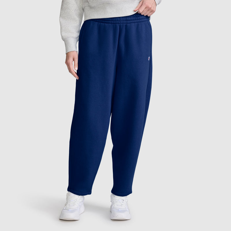 Dressed in Champion Barrel Leg Fleece Pant in navy blue, paired with a light gray sweatshirt and white sneakers, the person stands against a plain light gray background. The midweight fleece design of these Champion pants offers both comfort and style. Only the lower half of the body is shown.