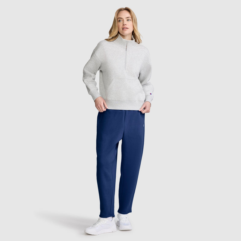 A person stands wearing a light gray pullover, Champion Barrel Leg Fleece Pant, and white sneakers, posing against a plain light gray background. The midweight fleece provides comfort and warmth to the relaxed look.