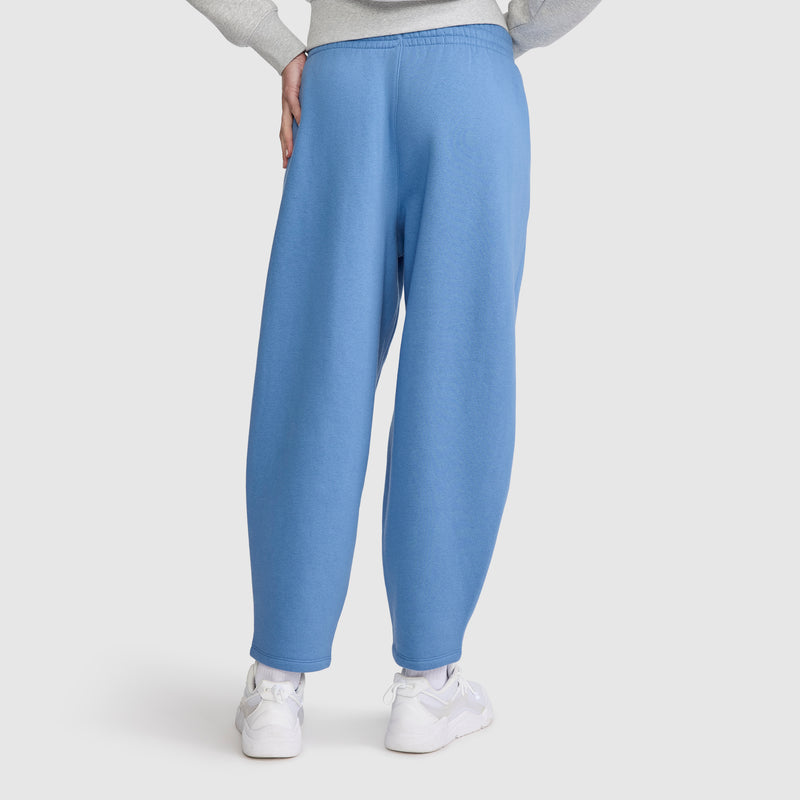 A person wearing Champion Barrel Leg Fleece Pant in light blue and white sneakers stands with one hand on their hip, facing away from the camera against a plain white background.