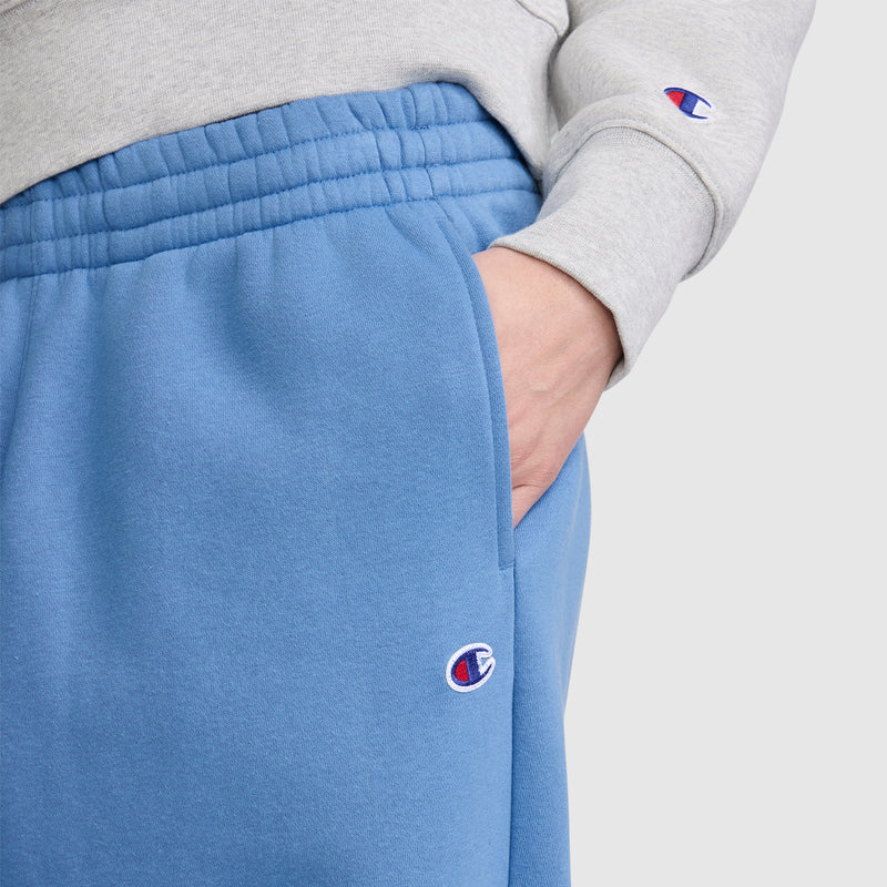 A person wearing a grey sweatshirt and blue Champion Barrel Leg Fleece Pants featuring contemporary sweats styling and subtle logos, with one hand in the pocket of the sweatpants.