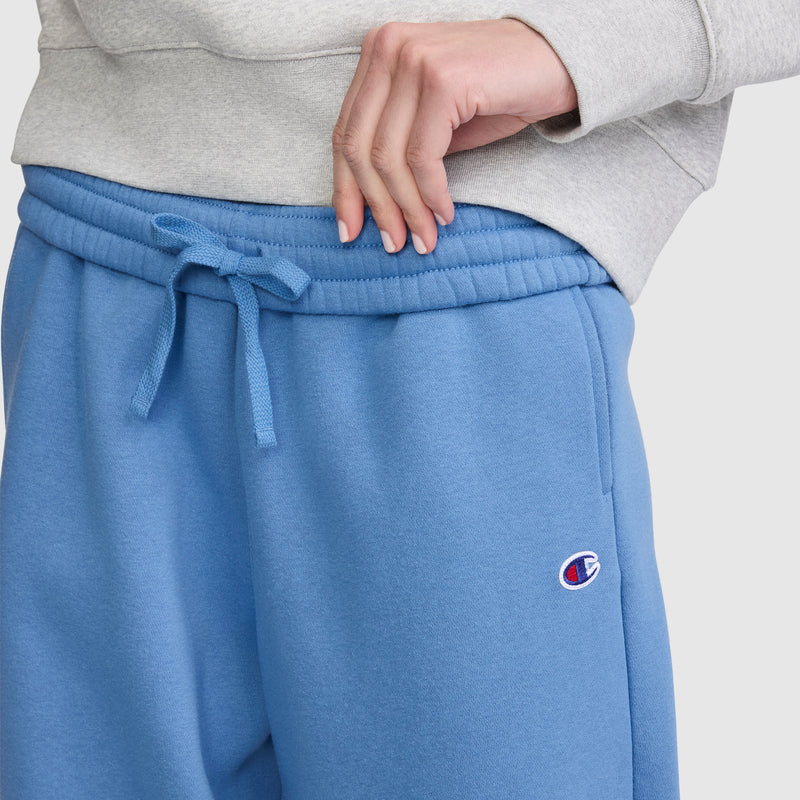 A person wearing the Champion Barrel Leg Fleece Pant in blue, featuring a relaxed fit with a drawstring waist and a small Champion logo on the left hip, paired with a light gray sweatshirt to showcase contemporary loungewear style.
