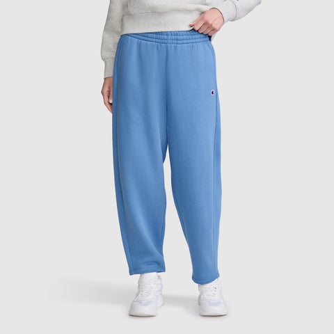 A person wearing contemporary sweats—specifically the Champion Barrel Leg Fleece Pant in blue, a light gray sweatshirt, and white sneakers—stands against a plain light background.