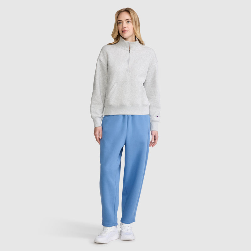 A woman with light-colored hair stands against a plain background, looking slightly to the side with a neutral expression. She is wearing a light gray half-zip sweatshirt, Champion Barrel Leg Fleece Pants in blue, and white sneakers.