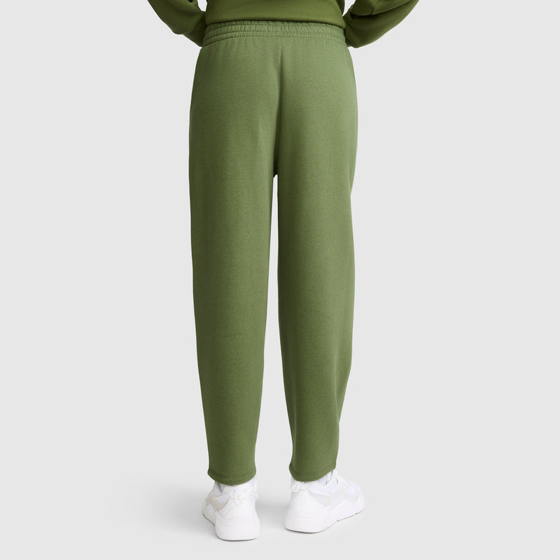 A person wearing Champion Barrel Leg Fleece Pants in olive green and white sneakers stands facing away with hands tucked in sweatshirt pockets, set against a plain light gray background.