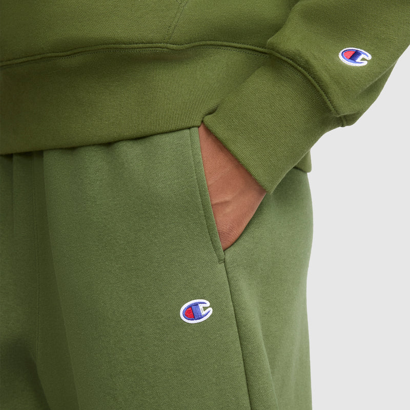 A person wearing an olive green sweatshirt and the Champion Barrel Leg Fleece Pant, featuring the embroidered Champion logo on the sleeve and pant pocket, stands with one hand in their pocket. The midweight fleece provides cozy comfort and a relaxed fit.