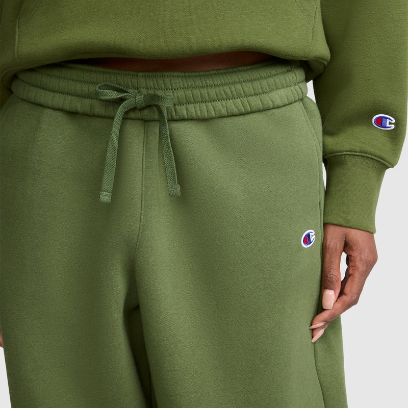 A person wears Champion Barrel Leg Fleece Pants in olive green along with a matching sweatshirt. One hand is visible by their side. Both garments display the Champion logo patches, and the midweight fleece fabric provides comfortable warmth.