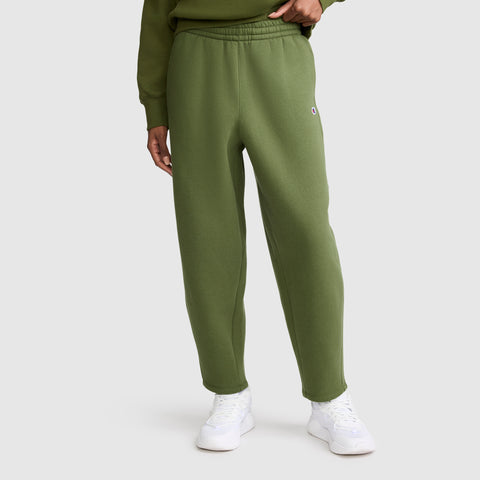 A person wearing Champion’s Barrel Leg Fleece Pant in olive green, paired with a matching midweight fleece sweatshirt and white sneakers, stands against a plain light gray background.