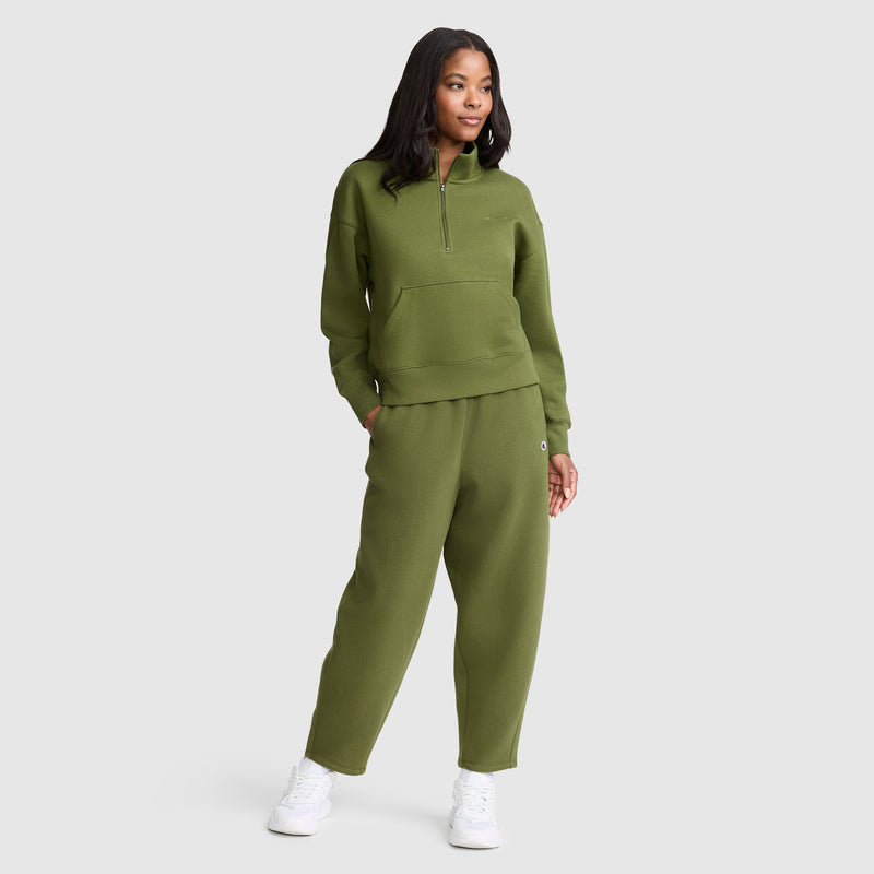 A woman stands wearing a matching olive green midweight fleece sweatshirt and Champion Barrel Leg Fleece Pants with white sneakers against a plain light gray background.