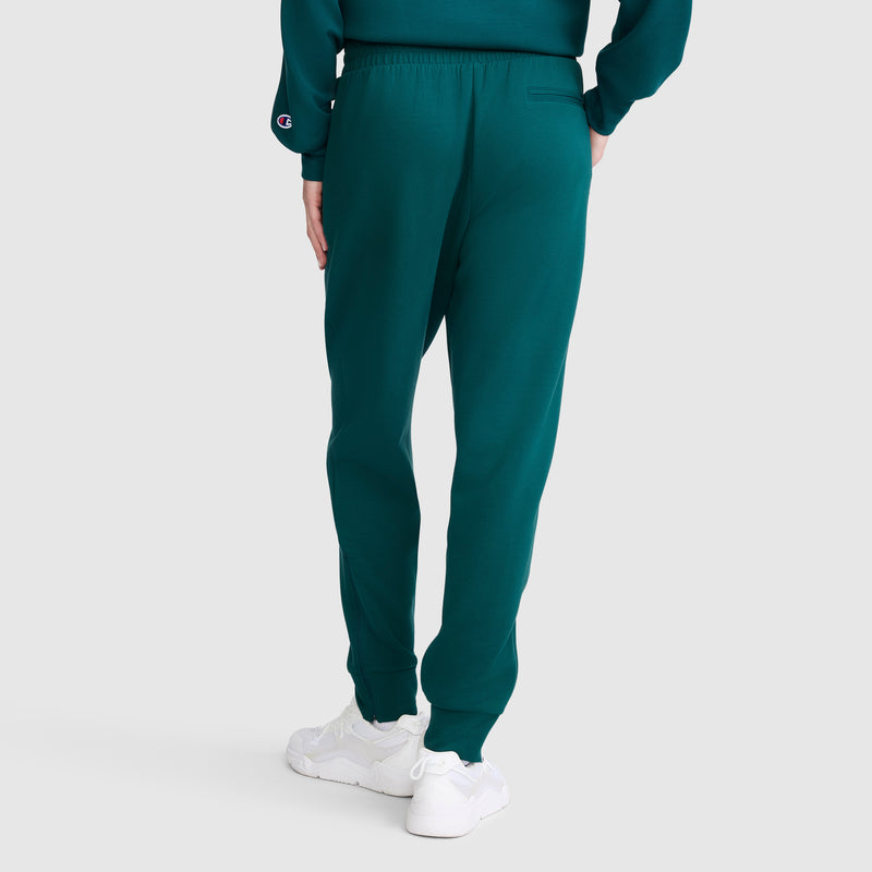 A person is seen from the back, standing against a plain light gray background, wearing teal Champion Taper Leg Jogger with Zippers pants paired with a matching teal sweatshirt and white sneakers.