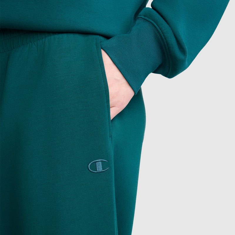 A person wearing teal Champion Taper Leg Jogger with Zippers and a matching sweatshirt has their hand in the pocket. The Champion logo is embroidered on the tapered leg of the jogger. The background is plain light gray.