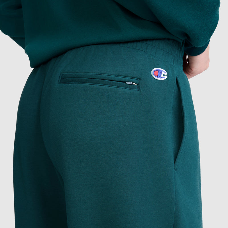 A close-up view shows a person wearing the Champion Taper Leg Jogger with Zippers, featuring a zippered back pocket and the signature Champion logo patch near the waistband. Their hand rests partially in the pocket, emphasizing the jogger’s comfortable fit and streamlined, tapered-leg design.