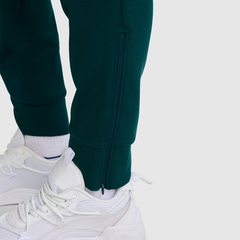 Close-up of a person wearing white sneakers, white socks, and the Champion Taper Leg Jogger with Zippers in dark green, featuring a visible zipper at the ankle. The background is plain and light-colored.