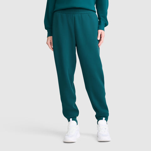 A person is wearing Champion Taper Leg Jogger with Zippers in teal and a matching top, paired with white sneakers. Only the lower half of the body is visible, standing against a plain light-gray background.