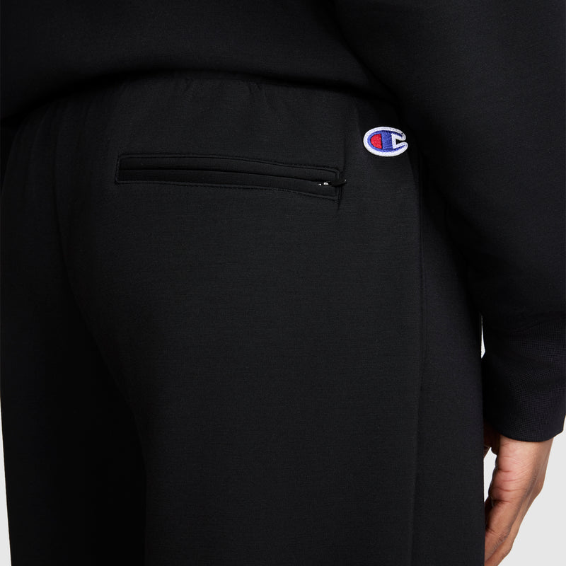 A close-up of a person wearing black Champion Taper Leg Jogger with Zippers, featuring a zippered back pocket and the Champion logo patch above it.