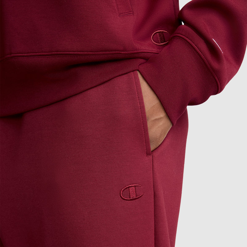 A person wearing a maroon Champion Taper Leg Jogger with Zippers tracksuit, their hand in the pocket. The Champion logo is visible on both the sweatshirt and the tapered leg jogger pants.
