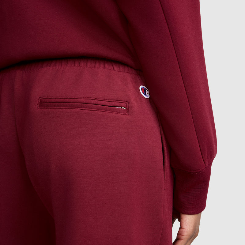 A person is wearing the Champion Taper Leg Jogger with Zippers in maroon, paired with a matching top. The joggers feature a small Champion logo patch near the right pocket, and their tapered leg design creates a flattering silhouette. Only the lower back and part of one arm are visible.