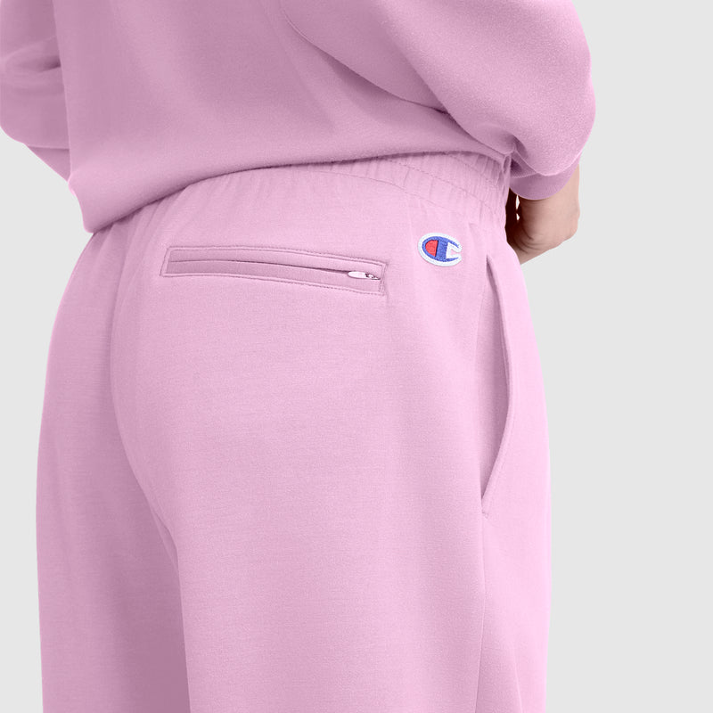 A person is shown from the back waist down wearing the Champion Taper Leg Jogger with Zippers in light pink. These joggers feature a tapered leg, a zippered back pocket, and the Champion logo on the right side. The background is plain and light gray.