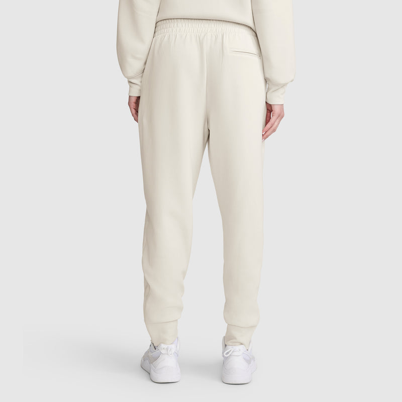 A person is seen from behind wearing cream-colored Champion Taper Leg Jogger with Zippers and a matching sweatshirt. The joggers feature a tapered leg, are made from a soft modal scuba blend, and have an elastic waistband. White sneakers complete the outfit, set against a plain light gray background.