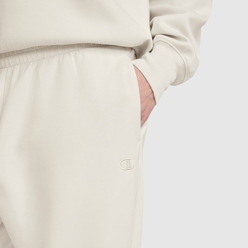 A person is wearing off-white Champion Taper Leg Jogger with Zippers and a matching sweatshirt, with one hand in the pocket. The Champion logo is embroidered on the jogger near the pocket, highlighting the signature style of Champion. The background is plain and light-colored.