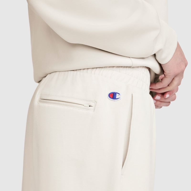 Wearing cream-colored Champion Taper Leg Jogger with Zippers and a matching top, a person stands with one hand on their hip, showcasing the Champion logo, side pocket zippers, and the sleek, tapered fit crafted from a soft modal scuba blend.