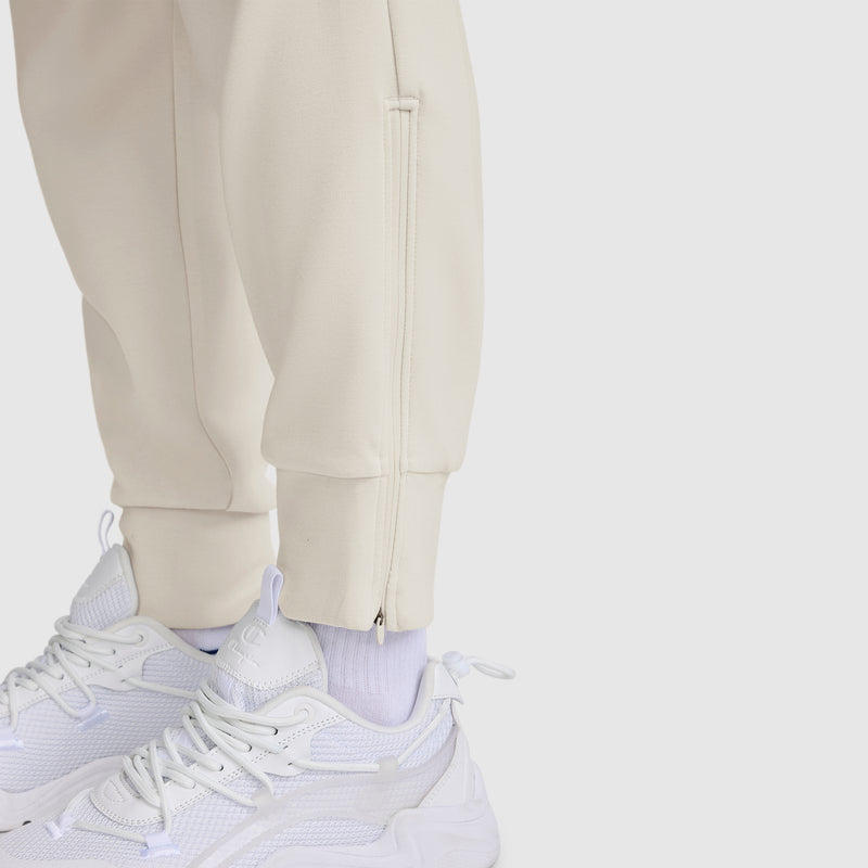 Close-up of a person wearing the Champion Taper Leg Jogger with Zippers in off-white, paired with white athletic sneakers and white socks, standing on a light gray surface. These joggers feature ankle zippers and are crafted from a comfortable modal scuba blend fabric for a modern, stylish look.