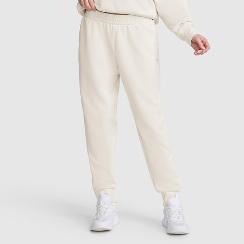 A person is shown from the waist down, wearing Champion Taper Leg Jogger with Zippers in light beige paired with a matching sweatshirt and white athletic shoes, standing against a plain light background. The joggers feature a modern tapered leg design.
