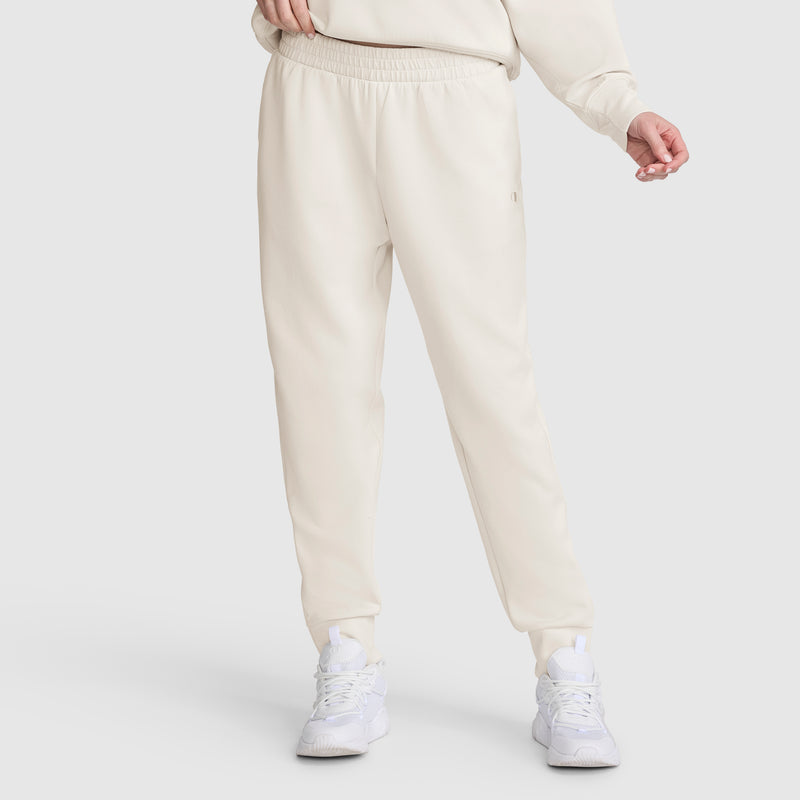 A person is shown from the waist down, wearing Champion Taper Leg Jogger with Zippers in light beige paired with a matching sweatshirt and white athletic shoes, standing against a plain light background. The joggers feature a modern tapered leg design.