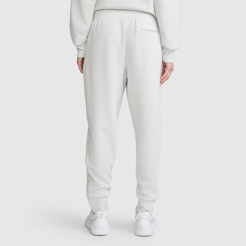 A person is seen from the back on a plain white background, wearing Champion Taper Leg Jogger with Zippers in white along with white sneakers. These women’s joggers by Champion feature an elastic waistband and a single back pocket on the right.