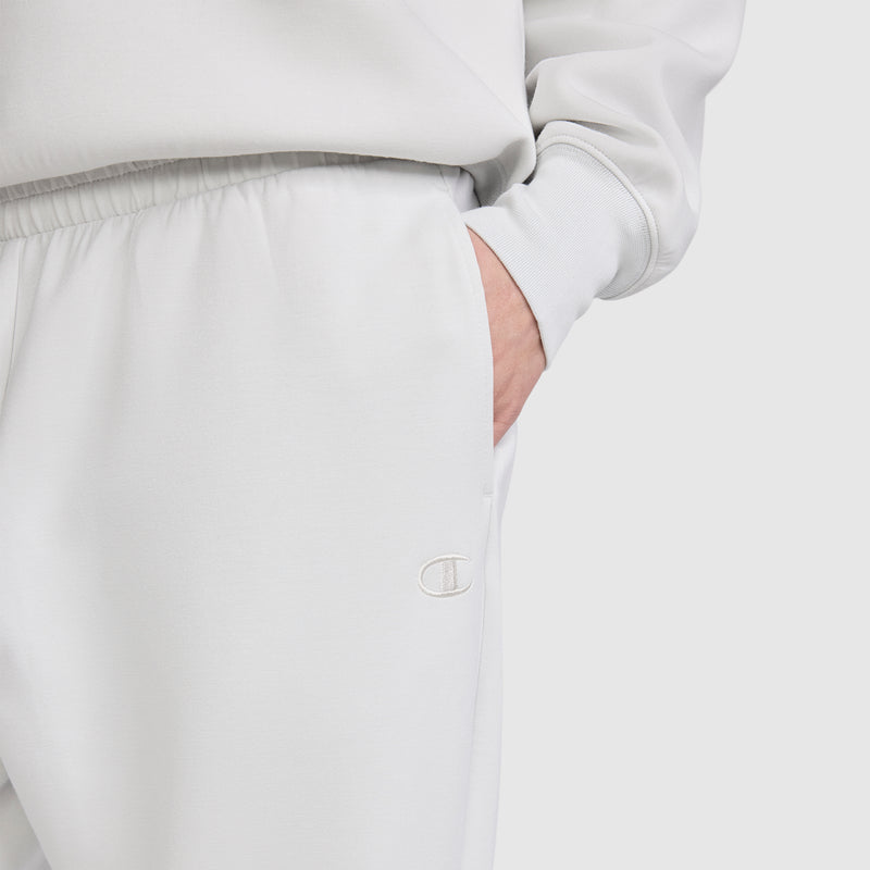 A person is wearing the Champion Taper Leg Jogger with Zippers in light gray, paired with a matching sweatshirt. Their right hand is in the pocket, and the Champion logo is visible on the joggers, which feature a tapered leg design. The background is plain and light-colored.