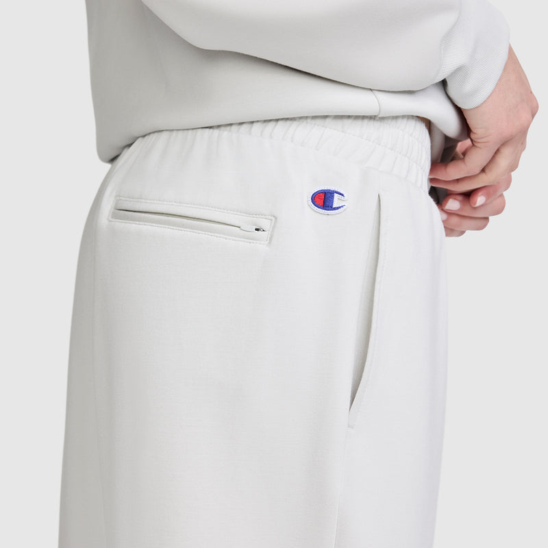 Close-up of a person wearing light gray Champion Taper Leg Jogger with Zippers, featuring an embroidered Champion logo above the pocket and a zippered back pocket. The tapered leg design is shown with hands resting on the hip, and a matching sweatshirt visible.