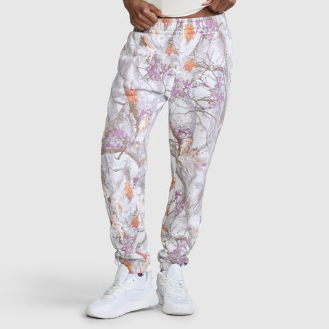 A model wears the Champion Powerblend Boyfriend Sweatpant with C Logo and an allover print featuring a gray, orange, and purple tree branch pattern reminiscent of camouflage. The jogger pants are styled with white sneakers and a white shirt, standing against a plain light gray background.
