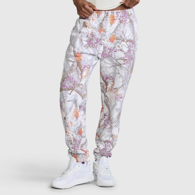 A model wears the Champion Powerblend Boyfriend Sweatpant with C Logo and an allover print featuring a gray, orange, and purple tree branch pattern reminiscent of camouflage. The jogger pants are styled with white sneakers and a white shirt, standing against a plain light gray background.
