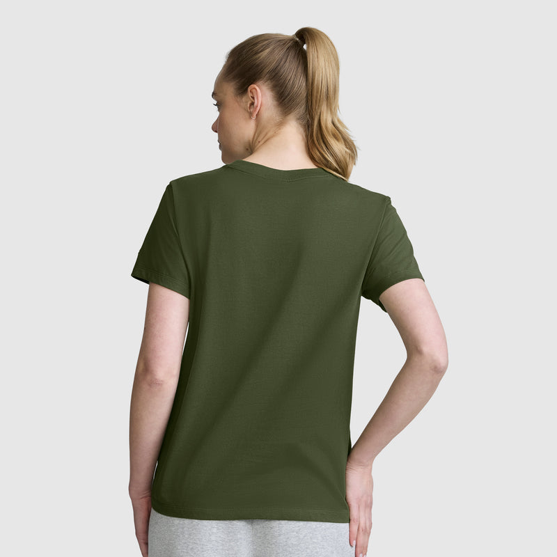 A woman with blonde hair in a ponytail stands with her back to the camera against a plain light background, wearing a Champion Mascot Graphic T-Shirt in olive green and gray pants.
