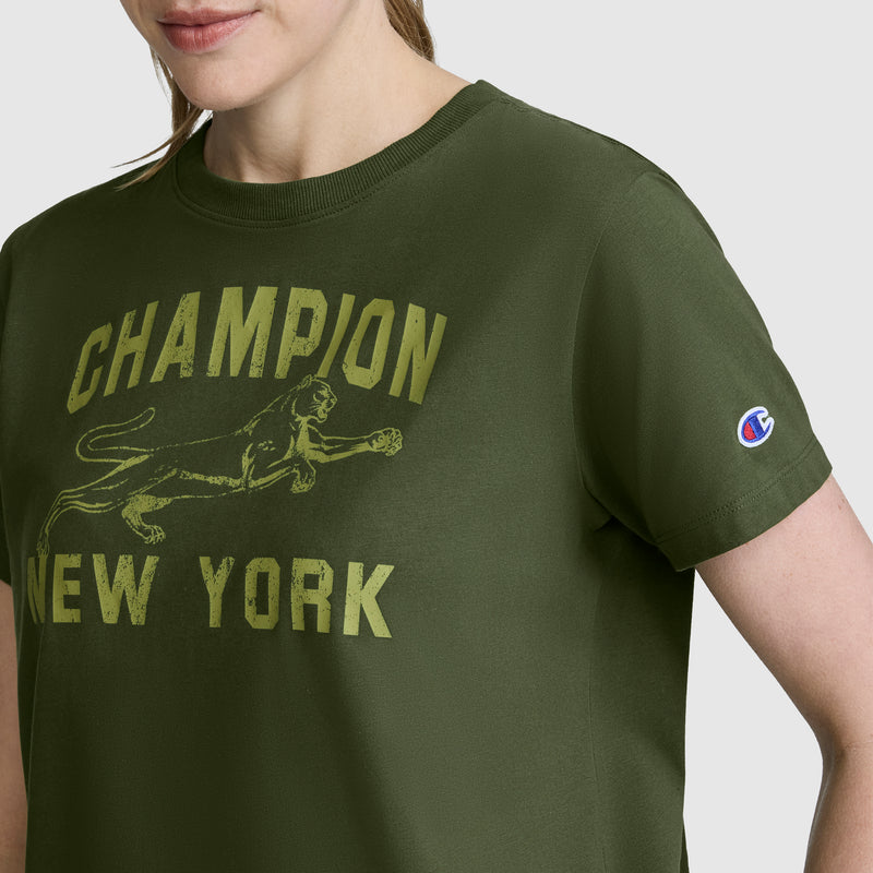 A woman wearing the Champion Mascot Graphic T-Shirt in olive green, featuring CHAMPION NEW YORK and a pouncing panther graphic on the front. This women’s tee includes the Champion logo on the sleeve, showcasing bold style from Champion.
