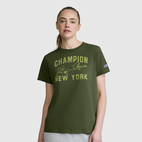 A woman stands against a plain light gray background wearing the Champion Mascot Graphic T-Shirt—an olive green women's tee featuring "CHAMPION NEW YORK" and a bold running dog graphic on the front.